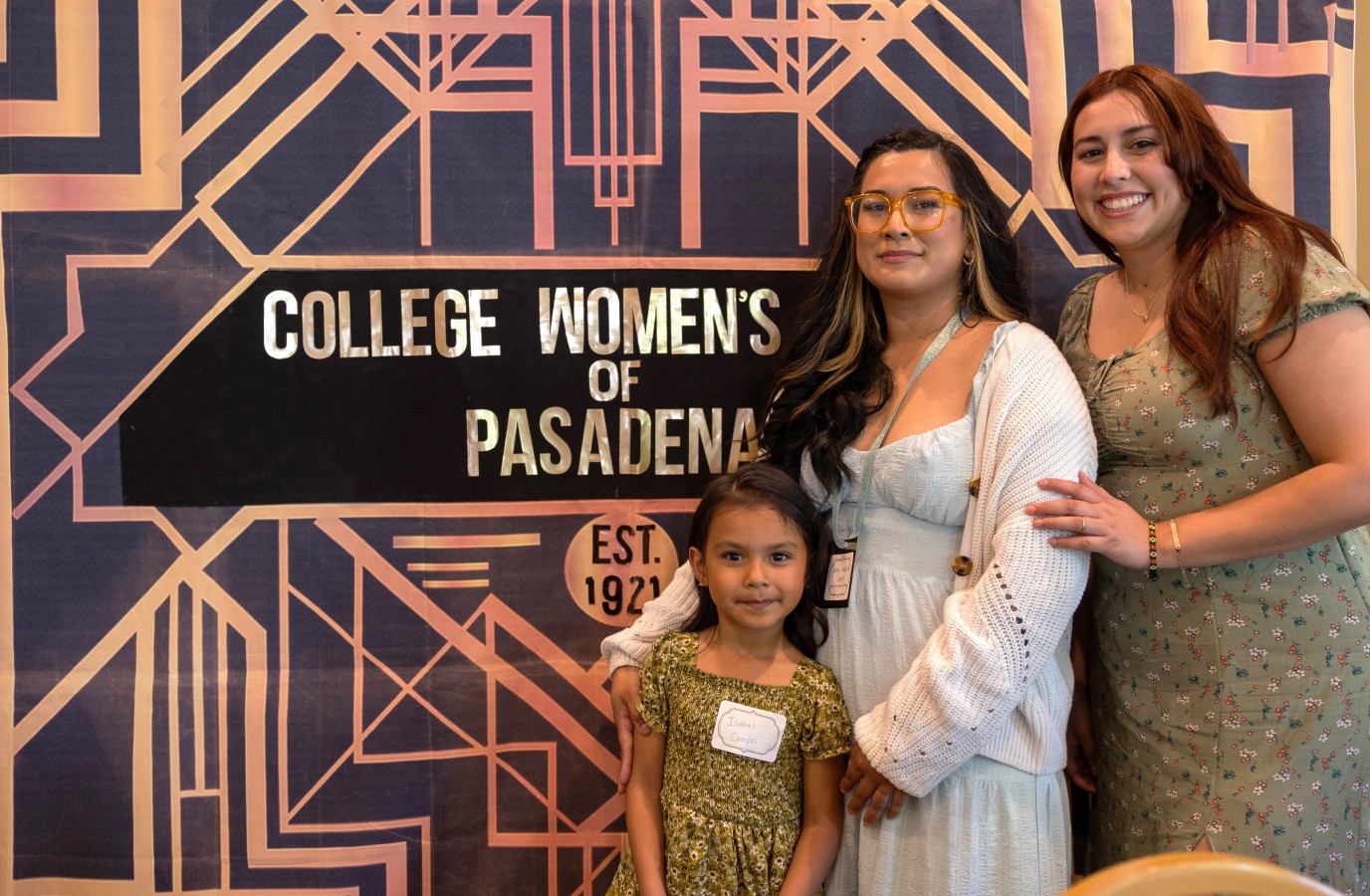Two women and a young girl taking a photo in front of the College Women of Pasadena (Great Gatsby) sign