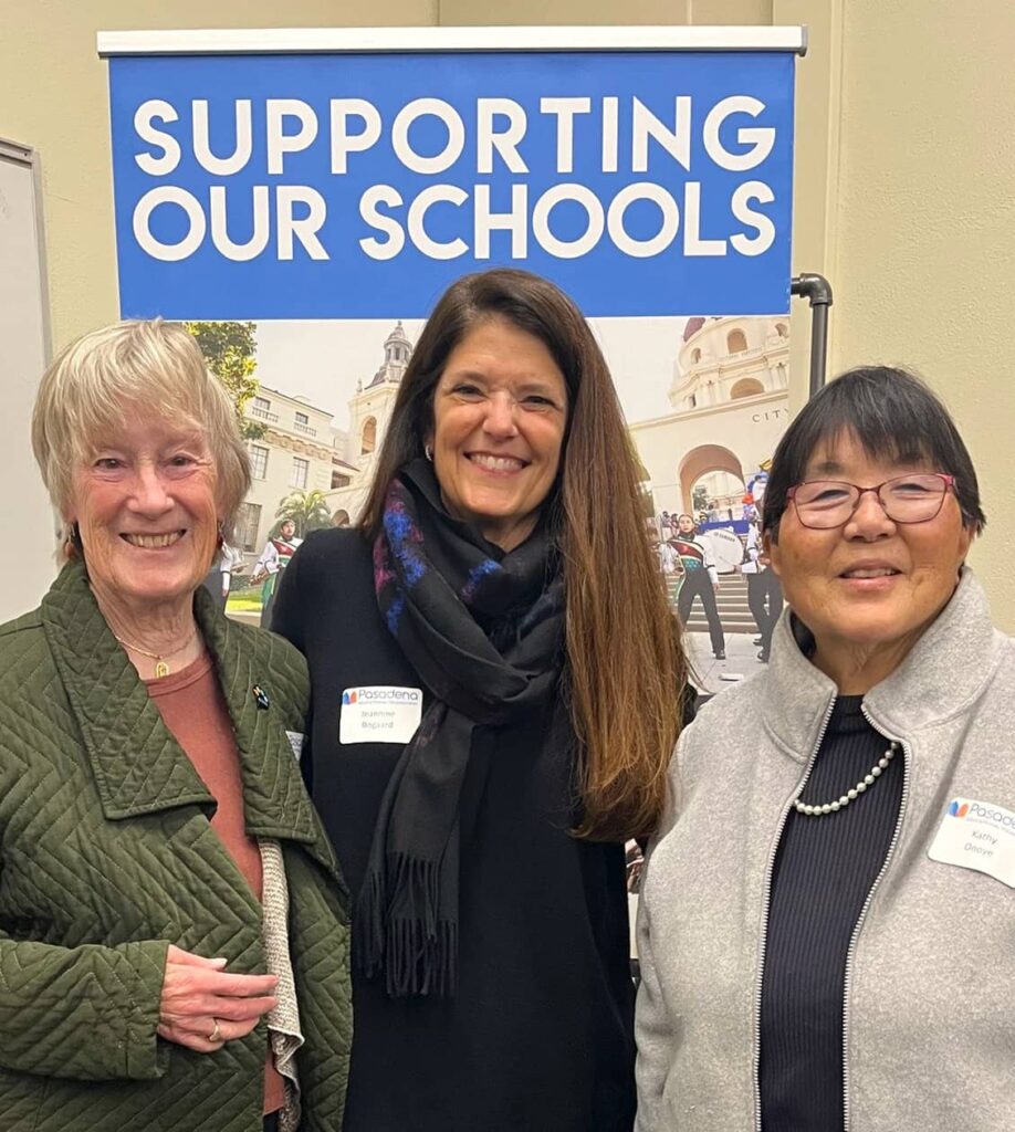 Three women smiling for a photo in front of a sign saying 'Supporting Our Schools'