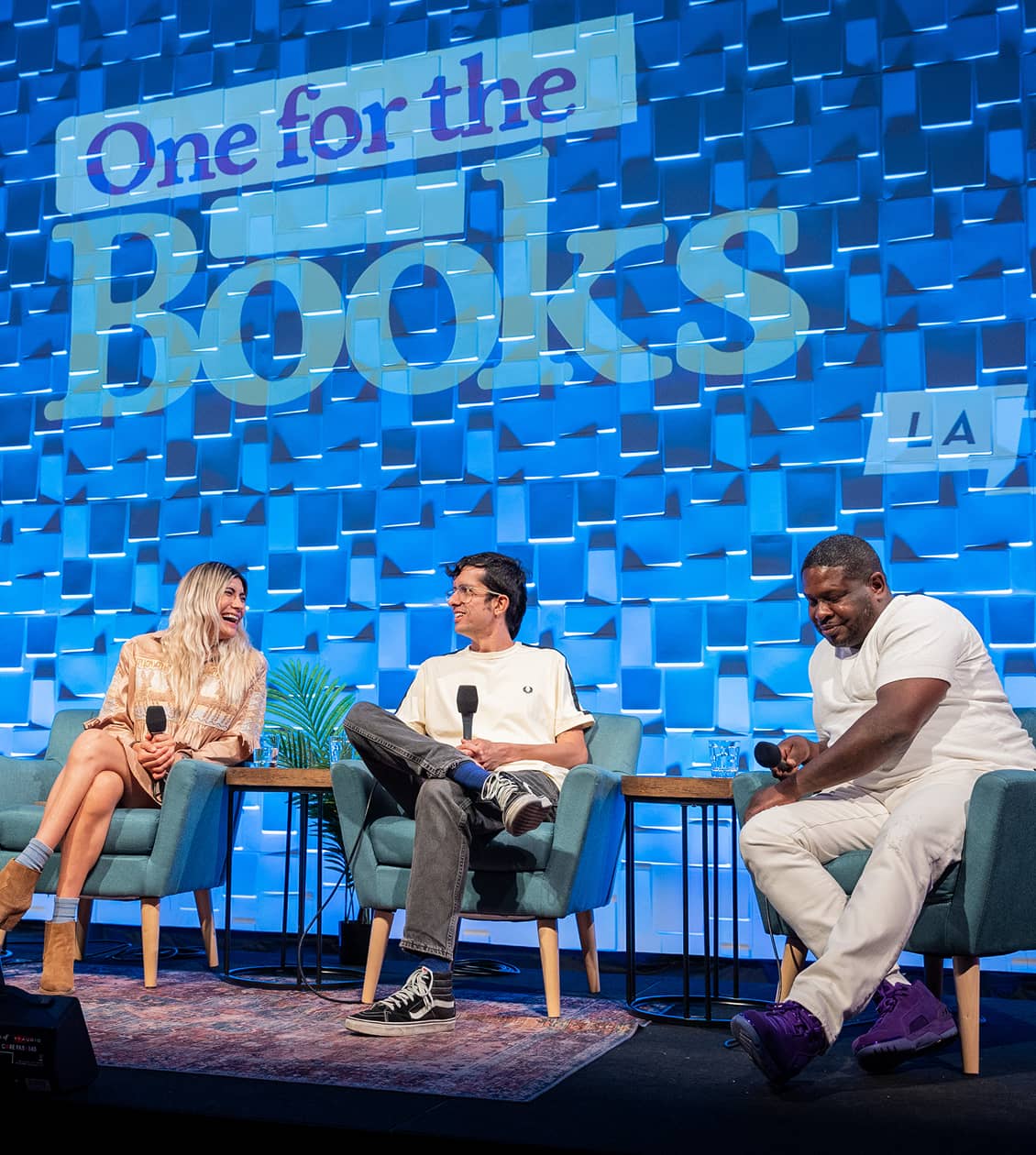 Two people sitting on stage discussing books with a projector banner stating 'One for the Books'