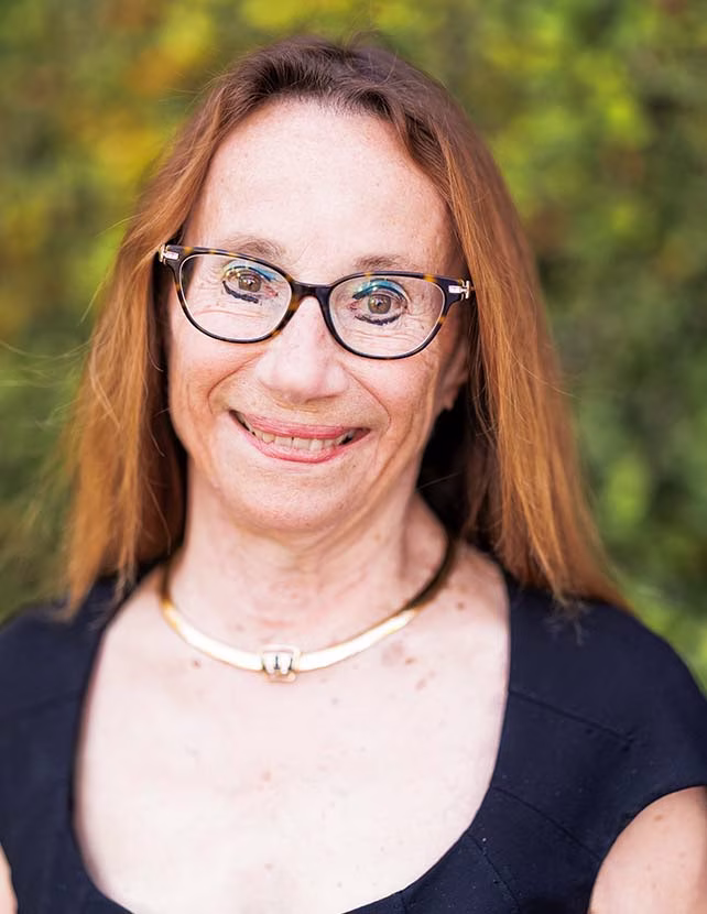 A woman in glasses and a black shirt with a gold necklace is smiling. Green leaf background.