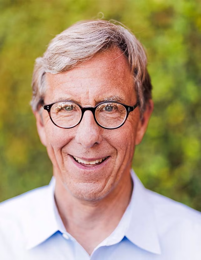 A man in gasses and a light blue button up shirt is smiling. Green leaf background.