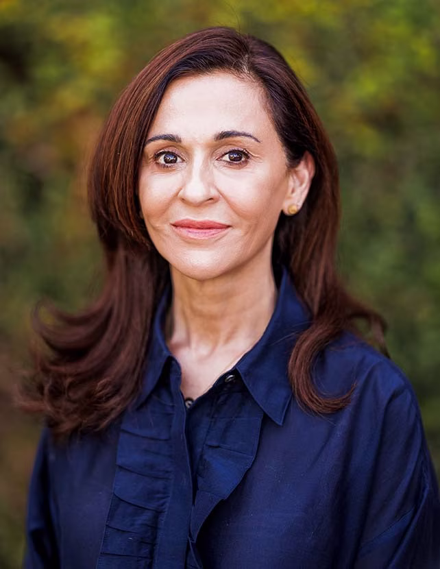 A woman in a blue button down shirt is smiling. Green leaf background.