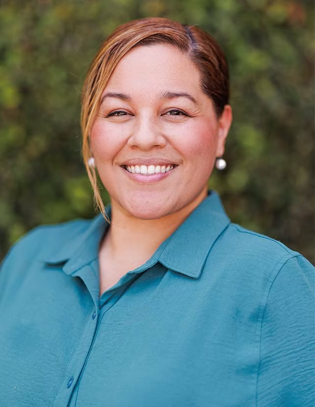 Woman in blue shirt is smiling. She is wearing earrings and has her hair in a bun. The background is green leaves.