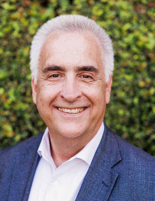 A man in a blue suit and white shirt is smiling. Green leaf background.