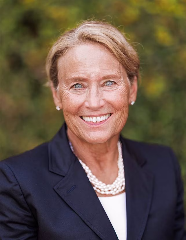 A woman is smiling in a back blazer. Green leaf background.