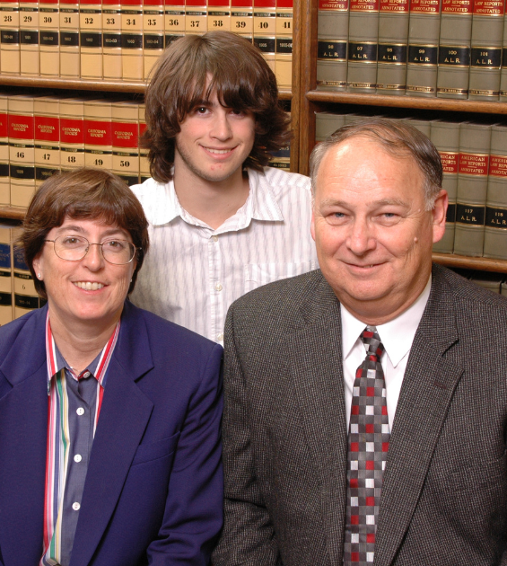 A middle age couple sits in a library with books surrounding them, behind them is a teen boy leaning into the photo