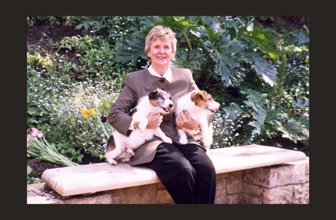 An older white woman wearing a professional suit sits on a park bench holding two dogs