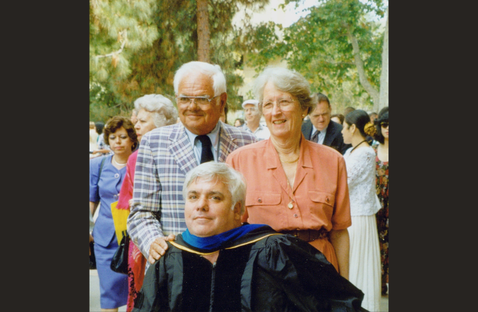 An older white couple stands behind a younger man wearing a graduation gown