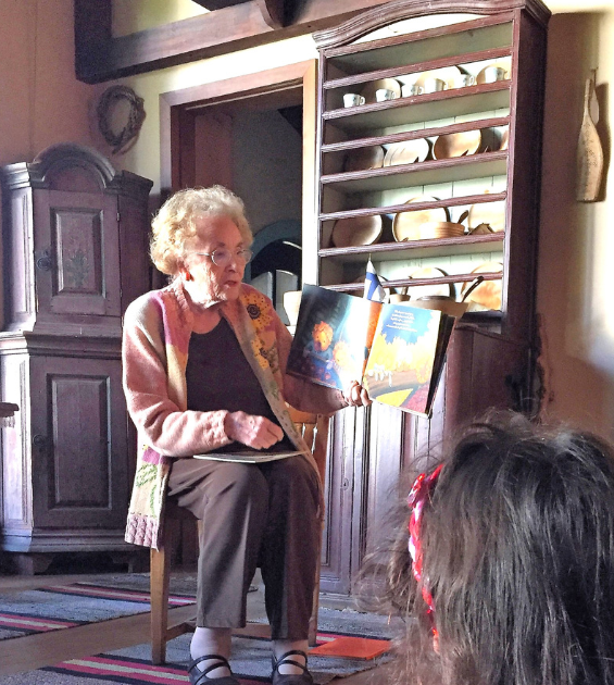 An elderly woman sits in a rocking chair holding a storybook while she reads, a child is in front of her listening