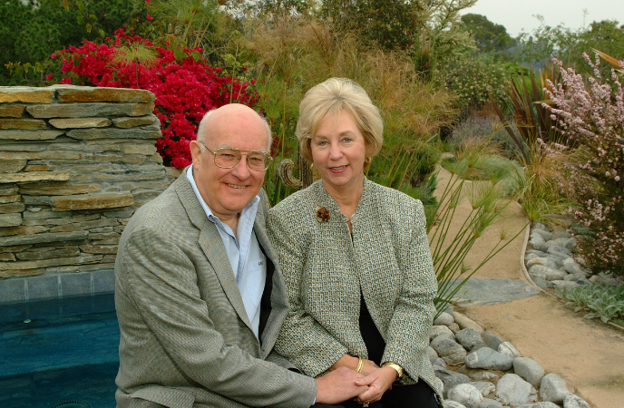 An older couple sits outside in a garden with a rock structure behind them