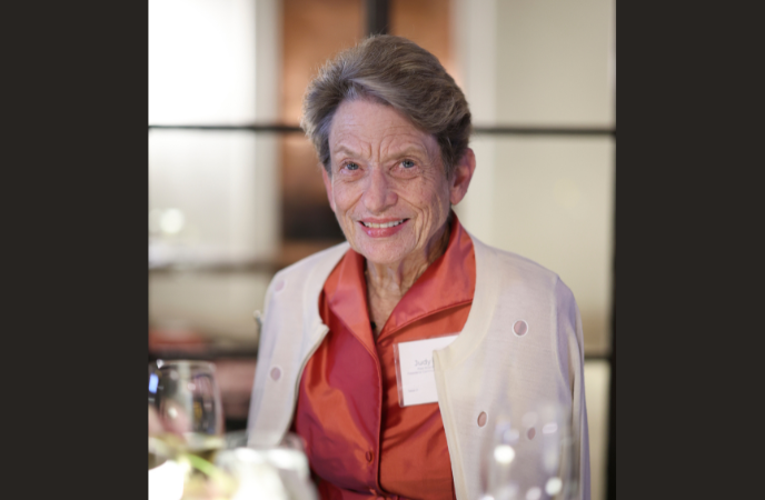 An elderly woman wearing a cardigan and silky red blouse sits at a dining table