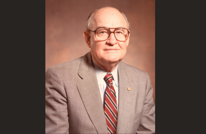 A vintage headshot of an older white male in a beige suit and brown tie