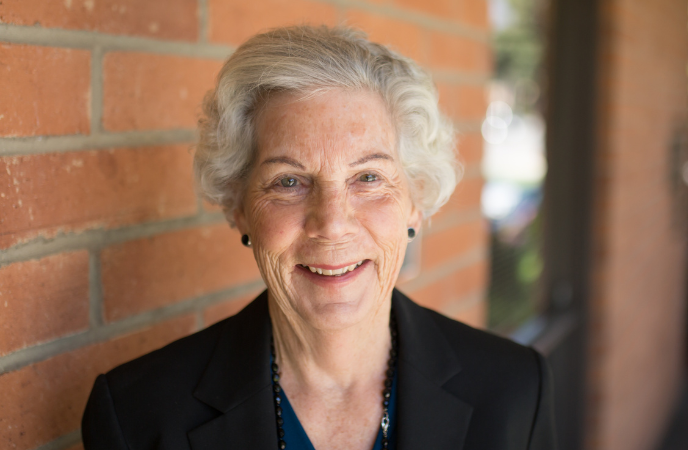 An elderly woman with short gray hair and wearing a black blouse stands outside in front of a brick wall