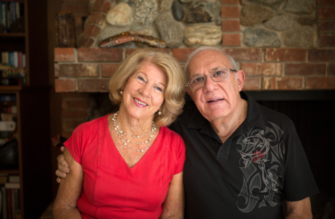 An older couple sit closely together in front of a fireplace