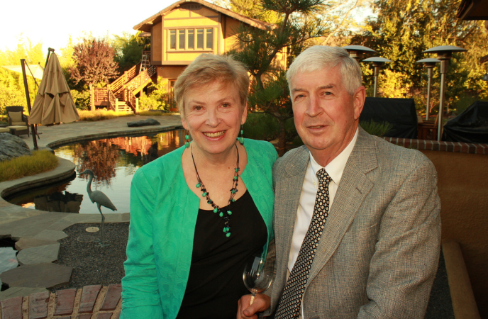 A couple in dress attire sits outdoors with a pond and house in the background