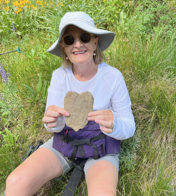 A woman wearing a hat sits outside in grass holding a heart-shaped rock