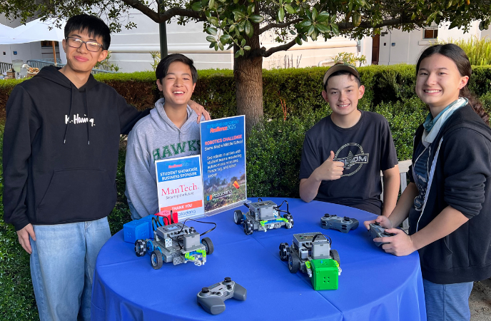 Four teens stand at a blue-tablecloth covered table wtih gadgets in front of them. A sign says