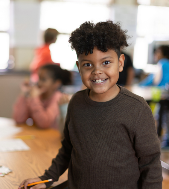 A young African American boy stands inside a classroom, hand resting on a table, facing camera. You can see other students behind him