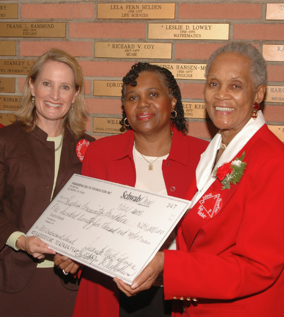 Two women in red suits hand a big check to a woman wearing a brown suit