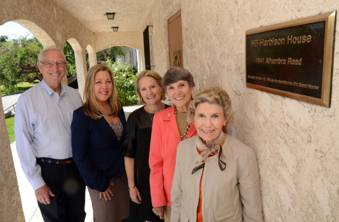 Five adults stand outside a building against a stucco wall and a wall plaque reads Hill Harbison House