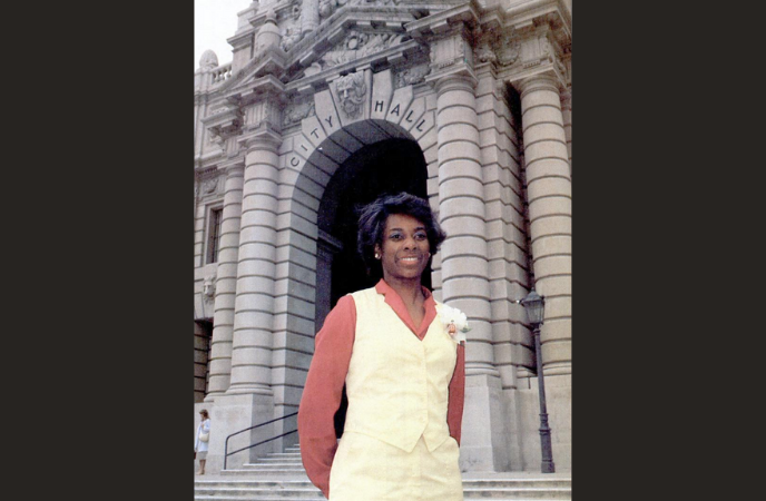 A woman in a 1970s style pantsuit stands on the steps of Pasadena City Hall