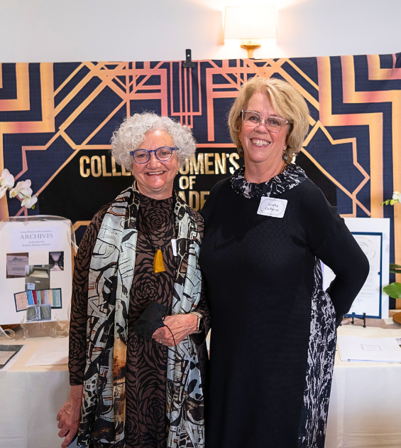 Two older women stand before a table with signage and banners