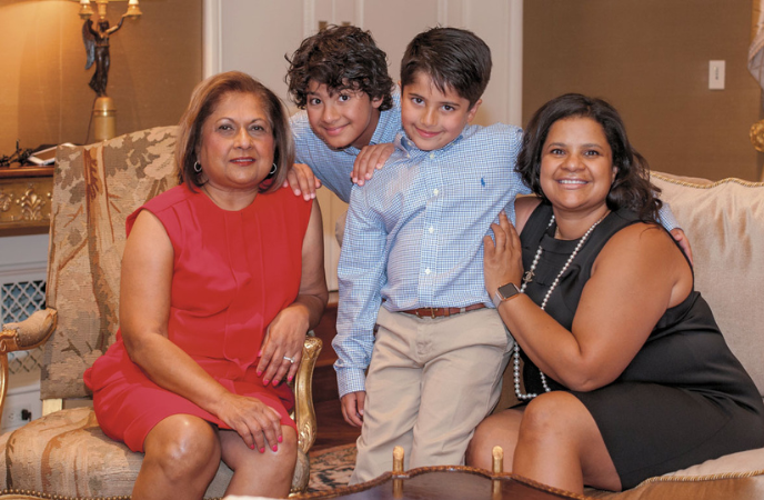An older woman in a red dress and a middle age woman in a black dress sit on a couch with two smiling young boys between them