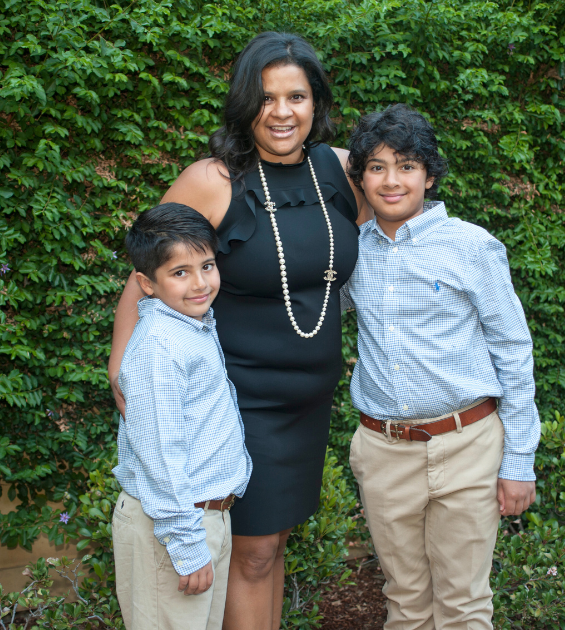 A middle age woman in a black dress stands outdoors, flanked by two boys each dressed in light blue shirts and khaki pants