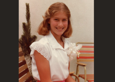A vintage photo shows a young girl in a white fluffy blouse and red skirt sitting outdoors on a patio