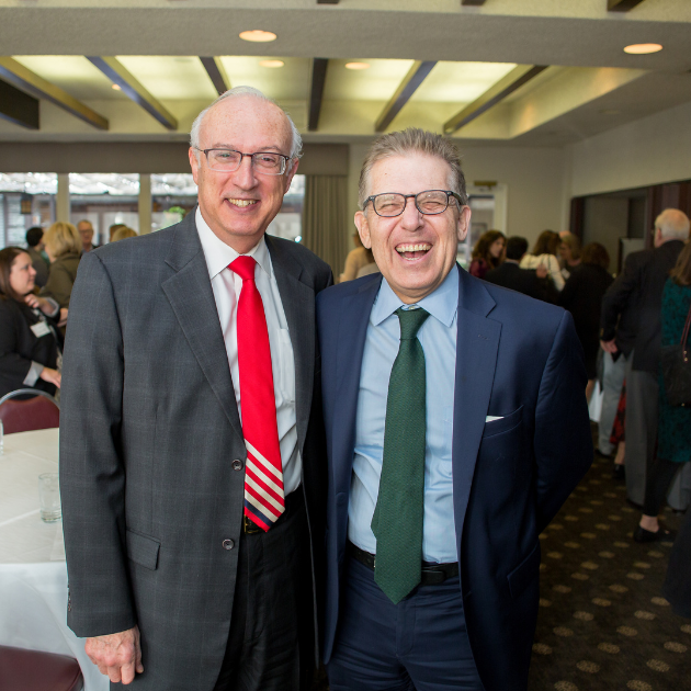 Two older men in suits stand together smiling and laughing at the camera