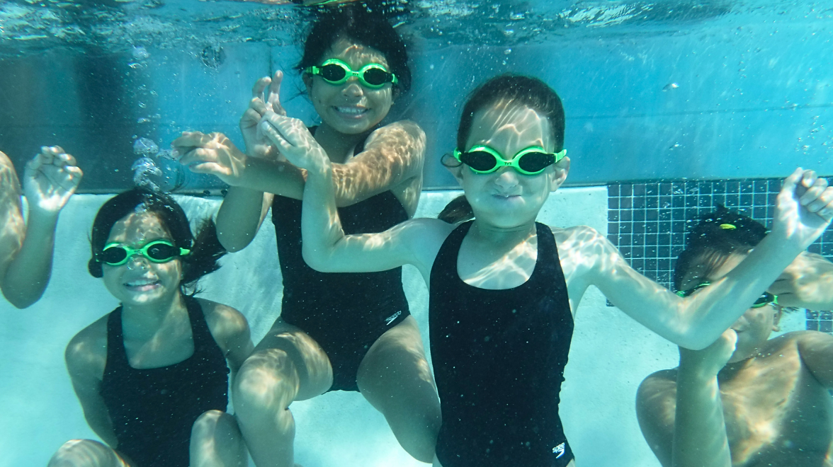 Four children wearing swimsuits and swim goggles are pictured underwater playing