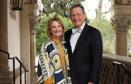 An older couple dressed in black tie attire stands under an arch