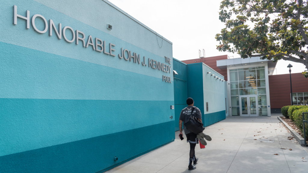 A man is shown from the back walking up a sidewalk to a doorway, next to him is a bright blue wall that reads honorable John Kennedy