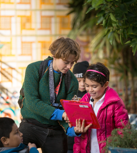 A young adult stands closely to a young girl showing her something on a clipboard