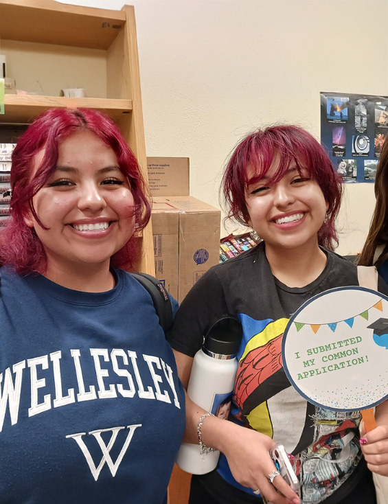 Two high school-age girls stand together. One wears a Wellesley sweatshirt and another holds up a paddle that reads "I submitted my common application"