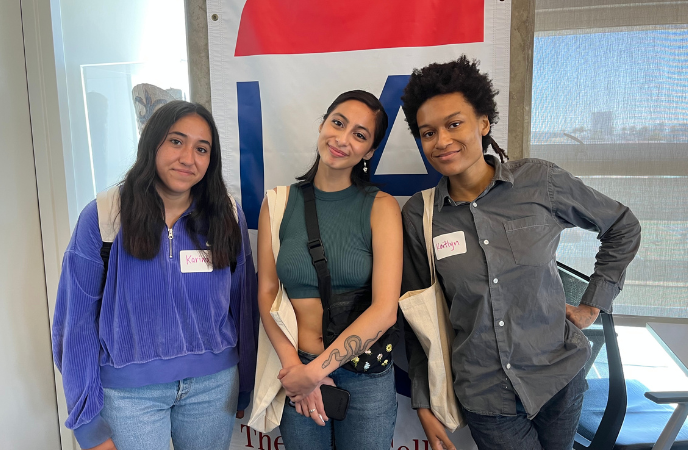 Three female college students stand together in front of windows and a banner