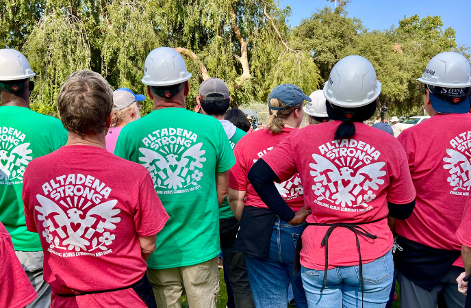 A group of people stand with their backs to the camera - they are all wearing hard hats and green/red shirts that read Altadena Strong