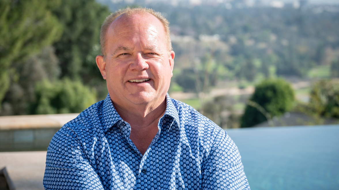 An older white gentleman in a blue shirt sits casually outdoors