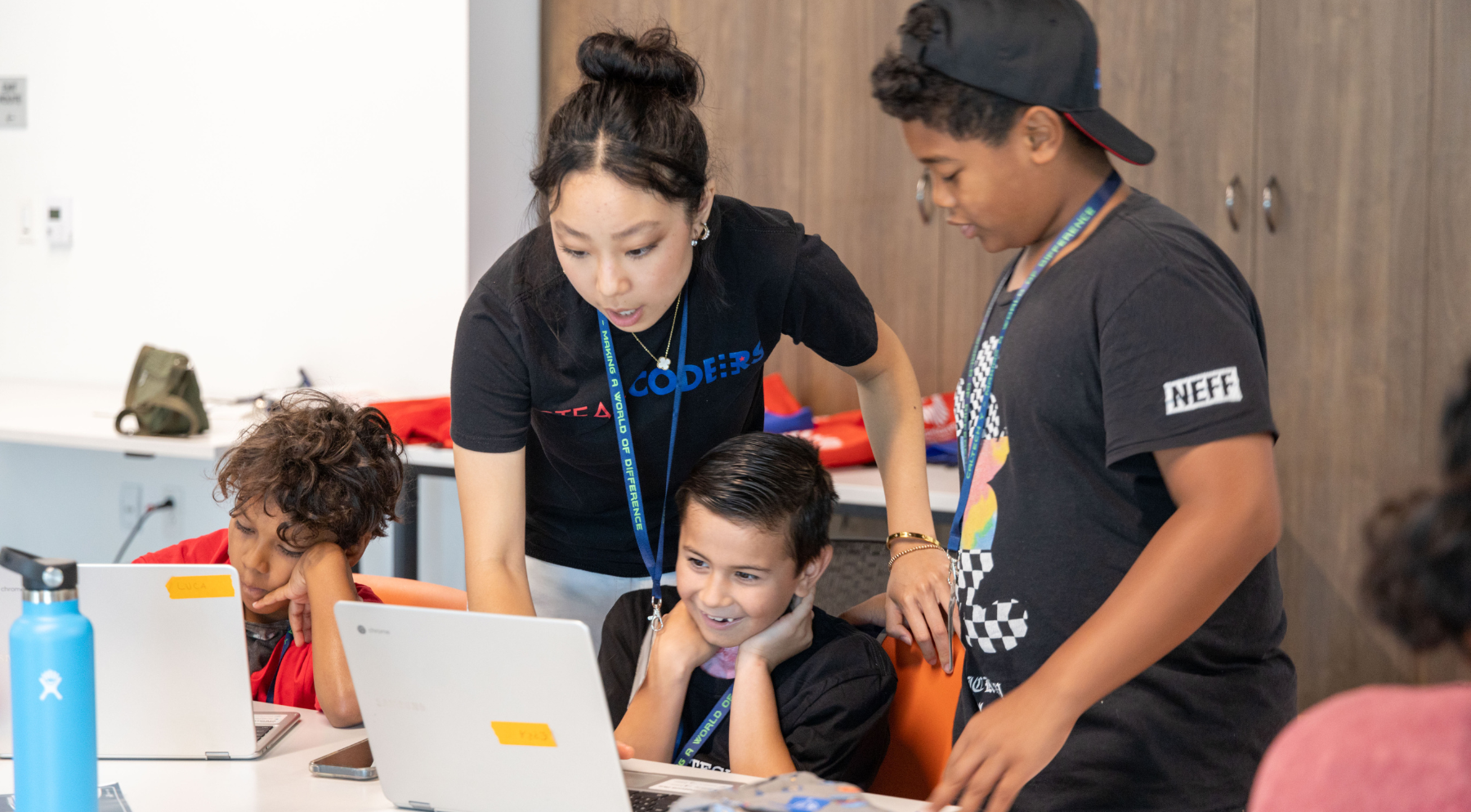 A young woman helps three teens and children with a laptop