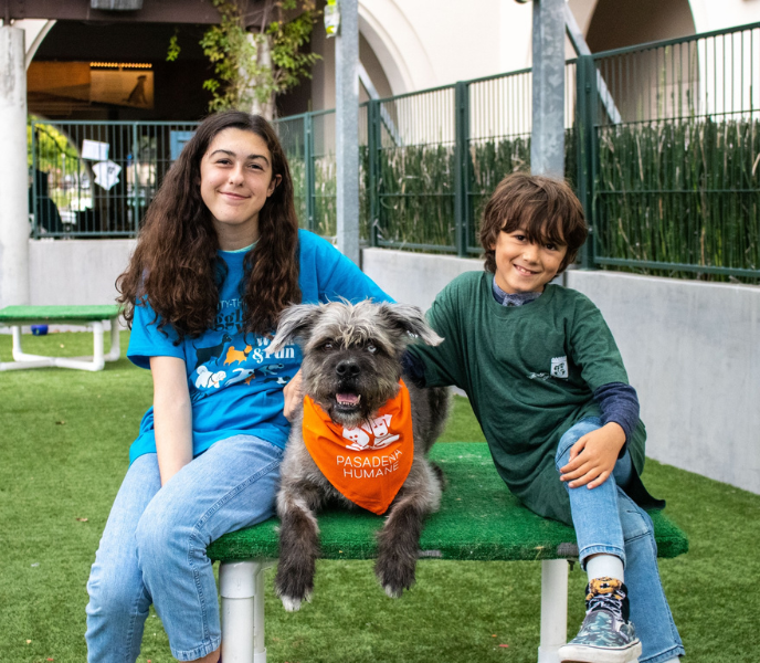 Two children - a boy and girl - sit in a grassy area with a dog in bewteen them. The dog wears an orange bandana that says Pasadena Humane