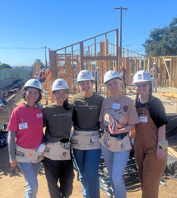 Five women wear hardhats and pose in front of a house being framed on a construction site