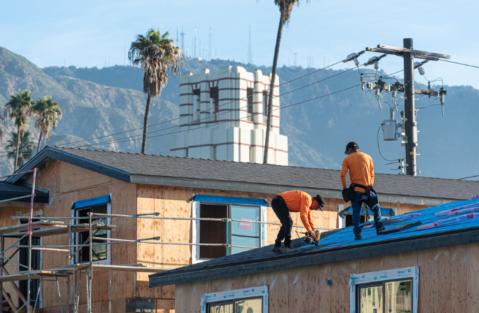 Two construction workers wearing bright orange shirts work atop a house undergoing construction. Altadena's signature Eliot Middle School tower and the San Gabriel Mountains are shown in background
