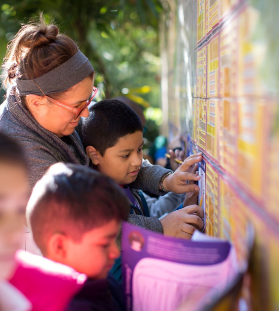 A woman is surrounded by a group of children outdoors, they appear to be touching and examining a tile wall