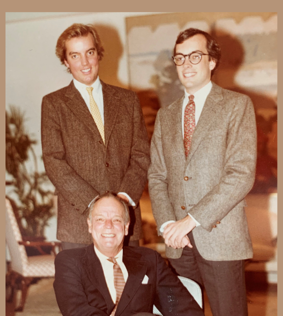 A vintage photo shows two young men standing and smiling behind an older man seated