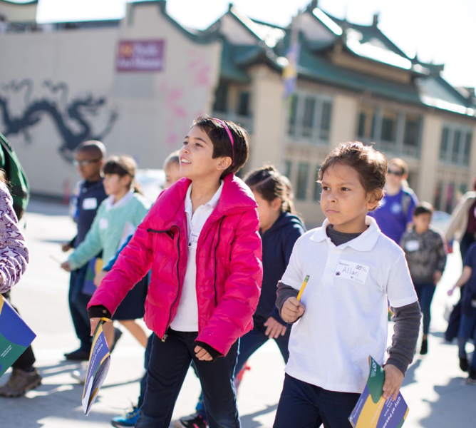 a group of children walk outdoors in Pasadena CA, in the forefront are a girl in a pink coat and a boy in a white shirt; both carry notebooks