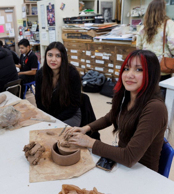 Two teen young women sit at a table inside an art studio; one has her hands on clay as she shapes a pot