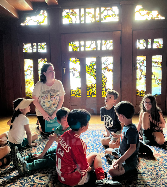A Group of young children sit with a teacher in front of brightly illuminate stained glass doors at the Gamble House in Pasadena CA
