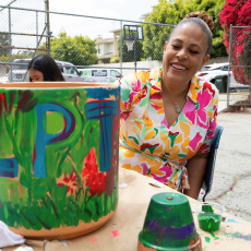 A woman in a bright floral dress sits back to admire a green garden pot she just painted