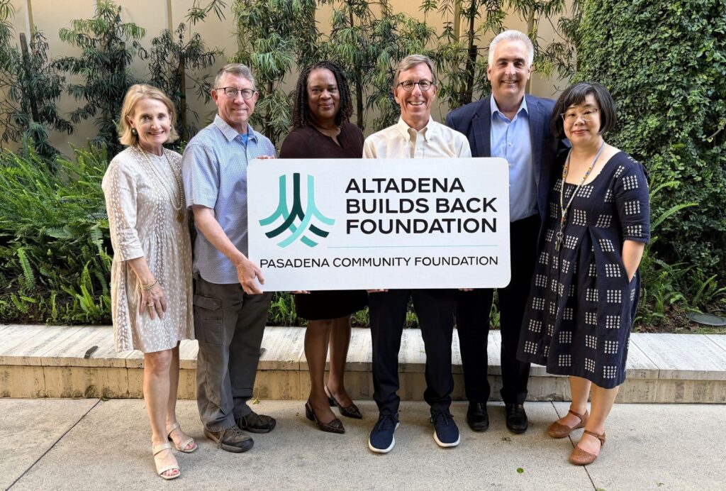 Six adults stand outdoors in a line, posing for camera while holding a sign that reads Altadena Builds Back Foundation and Pasadena Community Foundation