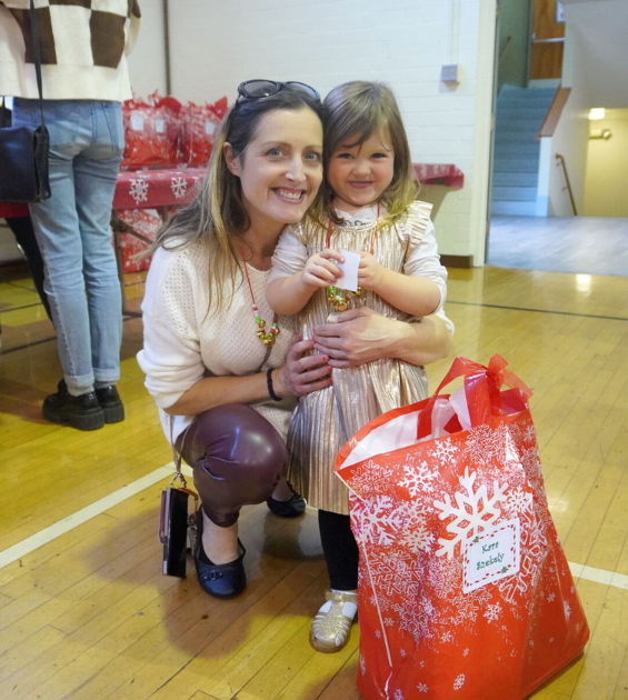 A young woman crouches down and has her arm around a little girl. In front of them is a festive holiday bag.